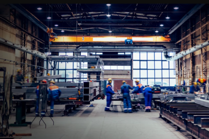Workers in high-visibility uniforms and safety helmets operating machinery and handling steel components inside a large industrial manufacturing facility.