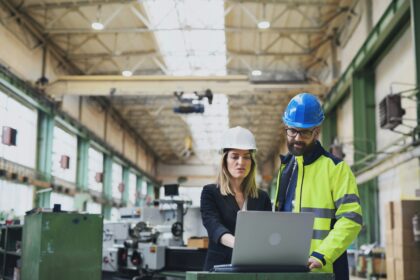 Two manufacturing professionals wearing safety helmets reviewing information on a laptop inside an industrial factory, with machinery in the background.